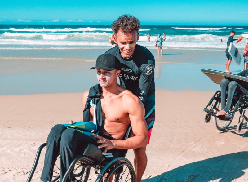 A person pushes a surfer in a wheelchair on the beach